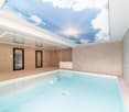 Modern indoor pool area with a large skylight showing a blue sky and clouds, featuring a rectangular swimming pool, a bathtub, and glass doors leading to another space.