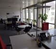 Office space with desks, chairs, screens, a round meeting table, potted plants, and office supplies.