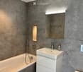 Modern bathroom with grey textured walls, a white bathtub, a white sink with a chrome faucet, a large mirror with built-in lighting, and a built-in shelf with ambient lighting.