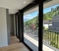 Modern hallway leading to a spacious balcony with wooden flooring, glass walls, and metal railings, overlooking a lush green landscape with trees and mountains.