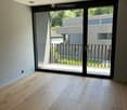 Empty room with light wooden flooring and large sliding glass doors leading to a balcony with a metal railing and greenery visible outside.