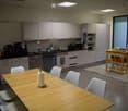 Modern kitchen with light grey cabinets, stainless steel appliances, a wooden dining table with white chairs, and a small cart with drinks and snacks.
