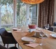 Dining area with a wooden table, chairs, wine glasses, plates, and a decorative bowl. Large windows with curtains and a hanging pendant light.