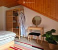Cozy bedroom with a wooden bed, a tall wooden wardrobe, a round mirror on a small table, a black stool, and a leafy plant in a woven basket.