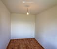 Empty room with wooden flooring and white walls, featuring a single hanging light fixture and electrical outlets.