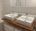 Modern bathroom with two white sinks on a wooden vanity, chrome faucets, a mirror, and a soap dispenser.