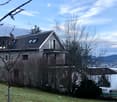 Two-story house with a modern design, featuring a sloped roof, multiple windows, and a balcony on the upper level. The house is surrounded by trees and offers a scenic view of the city and water in the background.