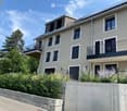 Modern two-story house with beige exterior, multiple windows, balconies, and a raised ground floor.