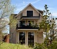 Two-story modern house with beige exterior, large windows, and balconies. The house features a triangular roof and is surrounded by greenery.