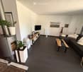 Modern living room with dark tiled flooring, a grey sectional sofa, a wooden coffee table, a television on a wooden stand, a fireplace with a plant on top, and a few decorative plants and artwork on the walls.
