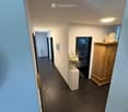 A narrow hallway with tiled flooring, light blue walls, and recessed ceiling lighting. There are several doors along the walls, a small white pedestal with a vase of white flowers, and a wooden cabinet with a decorative item on top.