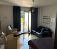 A modern, well-lit living space with a bed, sofa, coffee table, and chairs. Large windows and a sliding glass door lead to a balcony or terrace area.