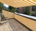 Balcony with tiled floor, yellow walls, a black folding chair, and a striped awning overhead. The view includes trees and neighboring buildings.