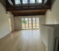 Empty room with wooden flooring, exposed wooden beams on the ceiling, large windows, and a sliding glass door leading to a balcony.