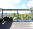 Modern rooftop terrace with a panoramic view of a green landscape and distant buildings. Features a clean, minimalist design with a metal railing, potted plants, and a spacious paved surface.