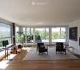 Bright, modern living room with large windows and a view, featuring a desk with a computer, a sofa, chairs, a coffee table, and a floor lamp.