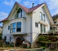 Two-story house with white exterior, red-tiled roof, multiple large windows, and a balcony on the upper level. The house is situated on a raised ground floor with a stone retaining wall and landscaped garden area.