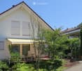 Two-story house with white exterior, red-tiled roof, and striped awning over the entrance. The front yard features a wooden deck with outdoor furniture, including chairs and a table, surrounded by greenery and plants.