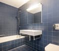 Modern bathroom with blue tiled walls and floor, featuring a bathtub, sink, mirror, and toilet.