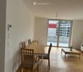 Bright, open-plan living and dining area with wooden flooring, a sofa, coffee table, dining table, and chairs. Large sliding glass doors lead to a balcony.
