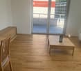 A modern, unfurnished living room with wooden flooring, a sliding glass door leading to a balcony, a wooden chair, a small table with a cup, and a wooden cabinet.