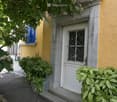 Entrance to a building with a white door, decorative window, potted plants, and a small bench nearby.