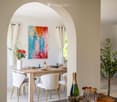 Elegant dining room with a wooden table, white chairs with gold legs, a vase with flowers, and a colorful abstract painting on the wall.