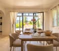 Elegant dining room with a wooden dining table set for a meal, surrounded by six upholstered chairs. Large windows with white curtains offer a scenic view of the garden. A vase with red flowers and a wine bottle are placed on the table.