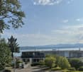 Modern two-story house with large windows and balconies, situated on a hillside overlooking a body of water and distant mountains.