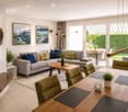 Modern living room with a grey sectional sofa, wooden coffee table, dining table with green chairs, floor lamp, fireplace, and artwork on the walls.