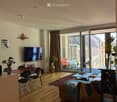 Modern living room with wooden flooring, large sliding glass doors leading to a balcony, sofa, coffee table, dining table with chairs, and a television mounted on the wall.