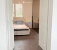 A clean, modern hallway with tiled flooring, white walls, and a ceiling light fixture. The hallway leads to a bedroom with a bed, pillows, and a small table with a lamp.