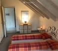 Cozy attic bedroom with slanted wooden ceiling, two single beds with red plaid bedding, nightstand with lamp, small table with chair, and wall-mounted picture frame.