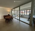 Bright, open-plan living and dining area with large windows and a view of the city. Features a grey sofa, wooden dining table with chairs, a cabinet, and a floor lamp.