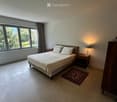 Spacious bedroom with a large bed, nightstand, lamp, and a small rug. Large windows with curtains allow natural light to fill the room.