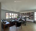 Bright living room with large windows, grey sofa, coffee table, bookshelf, and decorative plants.