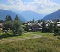 A scenic view of a village nestled in a valley with mountains in the background, featuring houses, trees, and a grassy field.
