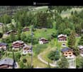 Aerial view of a picturesque village nestled in a lush, green valley surrounded by dense forests. The village features multiple wooden houses with balconies and terraces, scattered across rolling hills. The landscape includes well-maintained gardens, path