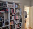 A cozy room with a large wooden bookshelf filled with books, a table lamp, and a cup on the shelf. An open door leads to another room with a visible refrigerator.
