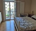 Cozy bedroom with a single bed, nightstand, radiator, and a cabinet. A balcony with a view of the street is accessible through large glass doors.