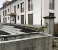 Modern residential building with white facade, multiple windows, balconies, and a decorative stone fountain in the foreground.