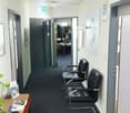 A clean, modern hallway with a dark blue carpet, white walls, and several doors. Two black leather chairs are positioned against the wall, and a small wooden table with brochures and a plant is visible on the left side.