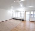 Empty room with white walls, wooden flooring, large windows with blinds, and a radiator. No furniture or equipment present.