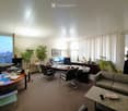 Office space with a sofa, coffee table, desk, office chairs, laptop, screen, and plants. Large windows offer a city view.
