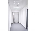 Modern, empty hallway with white walls, grey carpet, and recessed lighting. A single door is visible at the end of the corridor.