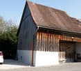 A rustic wooden barn with a tiled roof, white lower half, and a large wooden garage door. A white van is parked nearby.