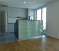 Modern kitchen with light grey cabinets, a central island with glass panels, and a refrigerator. The floor is wooden, and there are large windows letting in natural light.