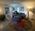 Modern living room with a grey sectional sofa, blue ottoman, floor lamp, and potted plants. The room features wooden flooring and a colorful round rug.