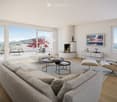 Modern living room with large windows offering a scenic mountain view, featuring a grey sofa, coffee table, chairs, a fireplace, and decorative items.