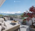 A spacious balcony with a panoramic mountain view, featuring a large sectional sofa, a coffee table, decorative lanterns, and potted plants including a bonsai tree and a large aloe vera plant.